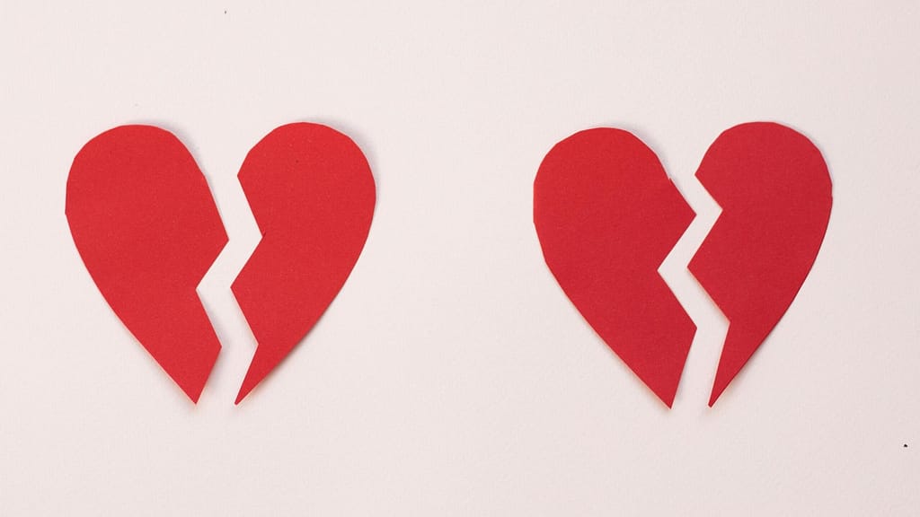 Two red broken hearts made out of construction paper sitting on a pale pink table symbolizing how to get over a breakup